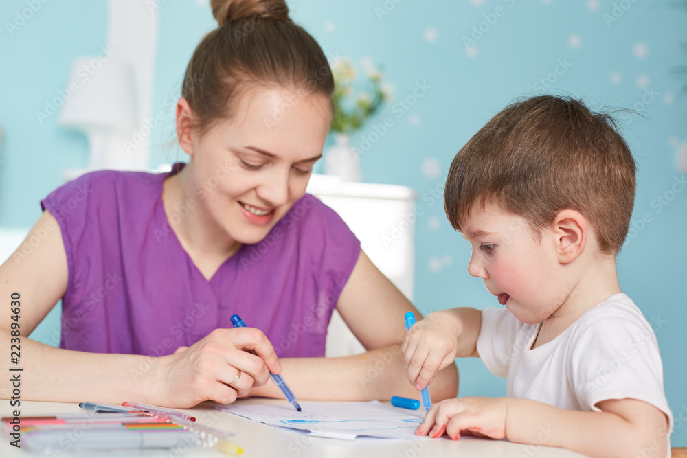 Photo of affectionate mother draws picture to her little son, spend ime together at home, have positive smiles, friendly relationships. Young mum takes care of child, gives educational lesson Photo of affectionate mother draws picture to her little son, spend ime together at home, have positive smiles, friendly relationships. Young mum takes care of child, gives educational lesson