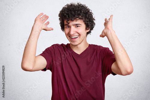 Happy teenager with curly hair, dressed in casual t shirt, gestures with hands, laughs at something, poses against white concrete wall. Delighted hipster expresses positive emotions and feelings