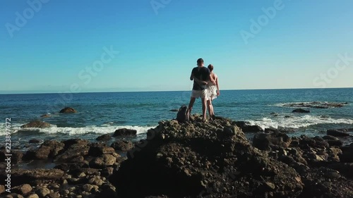 Young couple hugging, looking at the ocean. Woman raises her hand up. Back view. Gran Canaria.