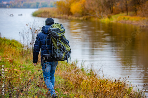 traveler with a backpack on his shoulders goes along the yellow grass along the bank near the river in the autumn