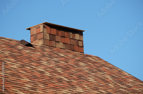 Part of brown roof of a house covered with soft shingles with short big smokestack close up under cloudless blue sky on sunny day