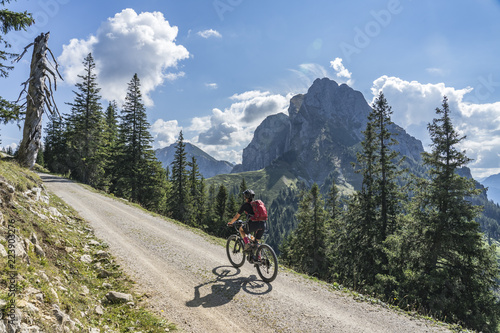 nice and active senior woman, riding her e-mountainbike in the Tannheim valley, Tirol, Austria with the village of Tannheim and famous summits Gimpel and Rote Flueh