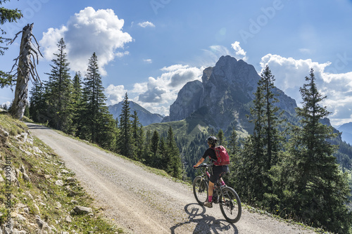 nice and active senior woman, riding her e-mountainbike in the Tannheim valley, Tirol, Austria with the village of Tannheim and famous summits Gimpel and Rote Flueh © Uwe