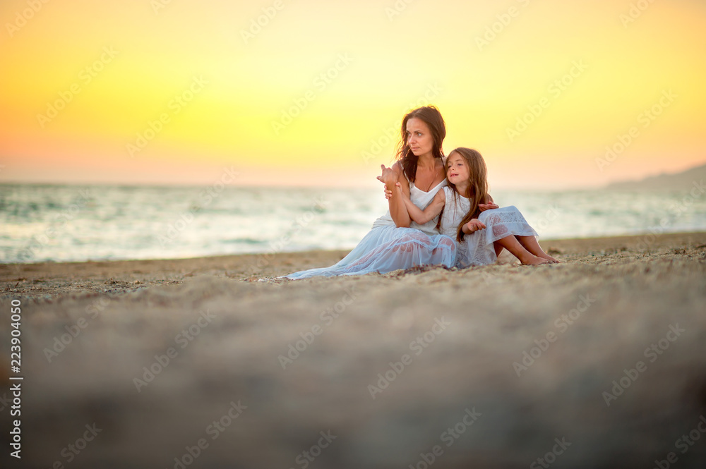 Mom with a little daughter on the beach at sunset, sitting on the sand and resting