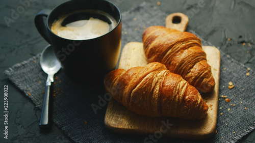 From above view of two fresh croissants and black mug with coffee placed on napkin on gray background of table