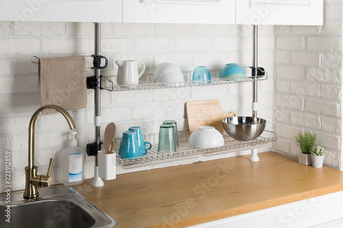 Clean dishes drying on metal dish rack on light background. Kitchen utensils and dishware on wooden shelf. Kitchen interior background.Text space.