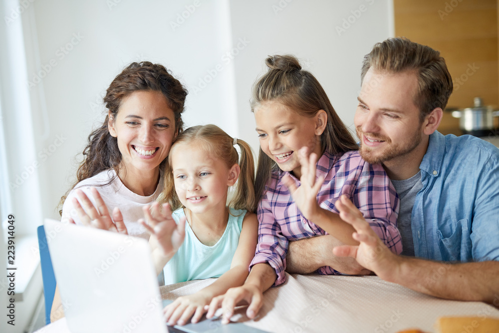 Young modern cheerful family of four sitting by table in front of laptop and talking through video-chat with their friends or relatives