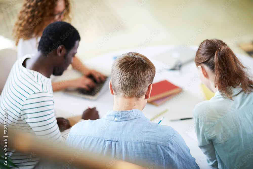 Group of young intercultural students in casualwear sitting by table and preparing for seminar individually Group of young intercultural students in casualwear sitting by table and preparing for seminar individually
