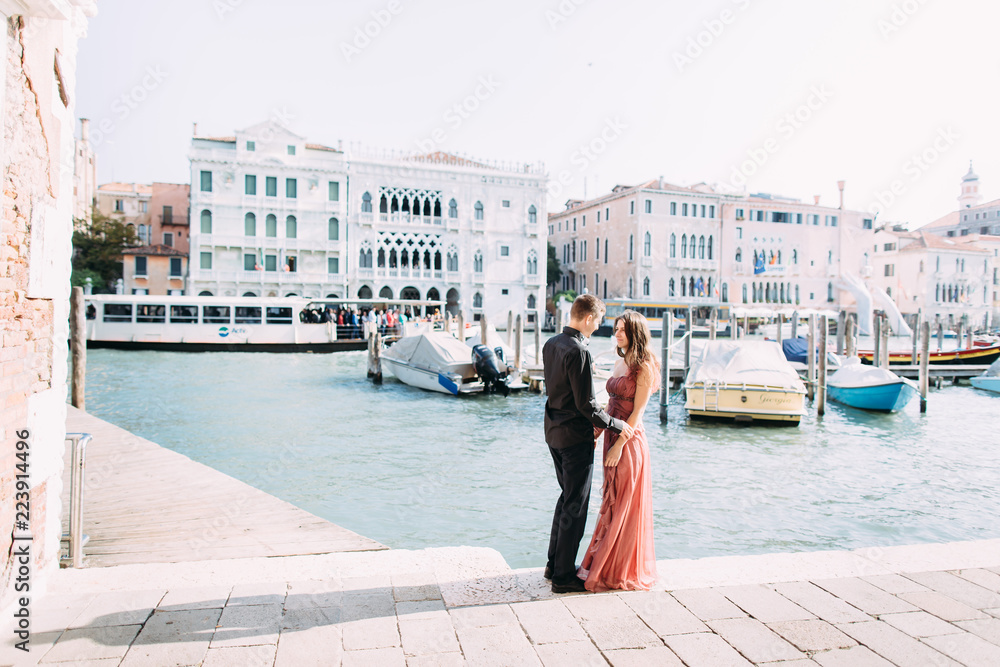 Romantic photo of hugging and kissing couple in Venice. Young couple, girl in pink dress and boy in black clothes standing on the background of wonderful Venice buildings. Italy, Europe.