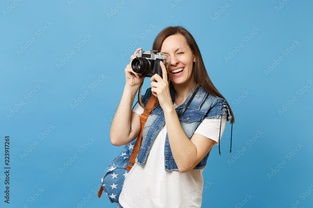 Portrait of young cheerful woman student in denim clothes with backpack taking pictures on retro vintage photo camera isolated on blue background. Education in college. Copy space for advertisement.