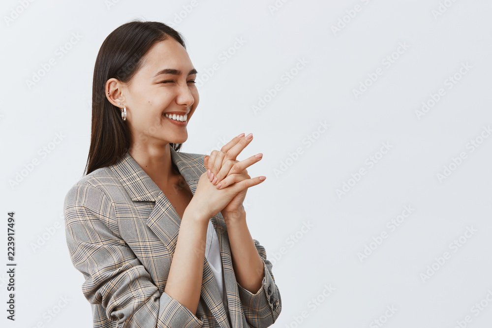 Studio shot of good-looking joyful woman laughing from joy and enthusiasm, standing half-turned over gray background, clasping hands together, looking right, cheering on b-day party Studio shot of good-looking joyful woman laughing from joy and enthusiasm, standing half-turned over gray background, clasping hands together, looking right, cheering on b-day party