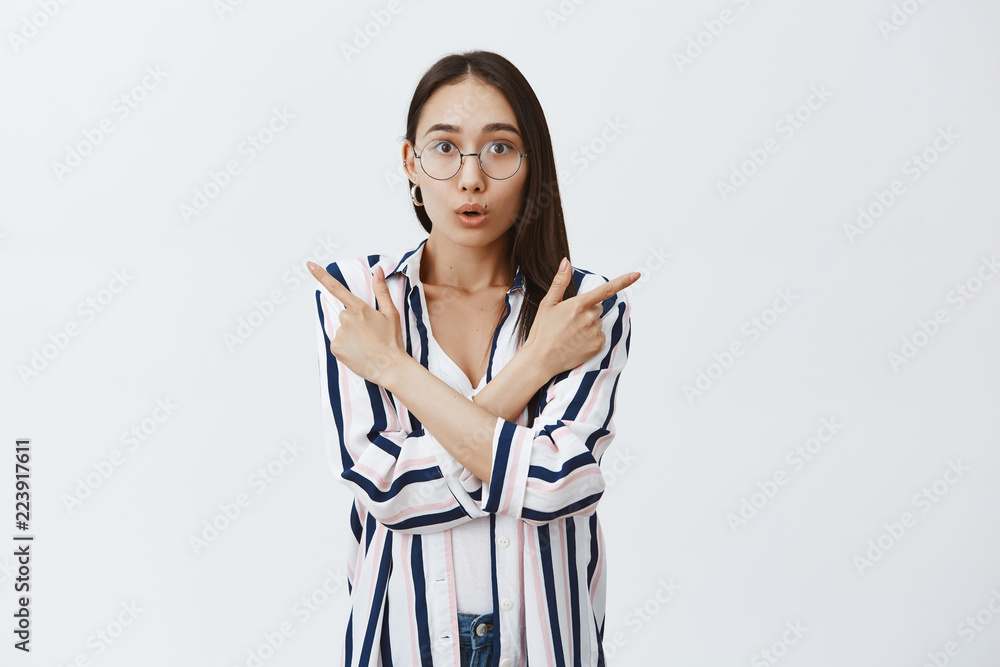 Indoor shot of amazed good-looking female entrepreneur in glasses and striped blouse, being fascinated and amazed on tourist travel, pointing right and left, folding lips from interest and thrill Indoor shot of amazed good-looking female entrepreneur in glasses and striped blouse, being fascinated and amazed on tourist travel, pointing right and left, folding lips from interest and thrill