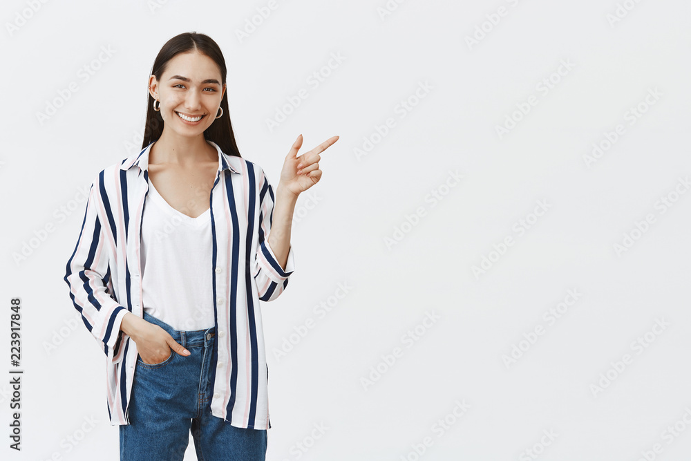 Shall we visit that place. Portrait of friendly good-looking and stylish female coworker in striped blouse and jeans, smiling broadly while holding hand in pocket and pointing right with forefinger Shall we visit that place. Portrait of friendly good-looking and stylish female coworker in striped blouse and jeans, smiling broadly while holding hand in pocket and pointing right with forefinger