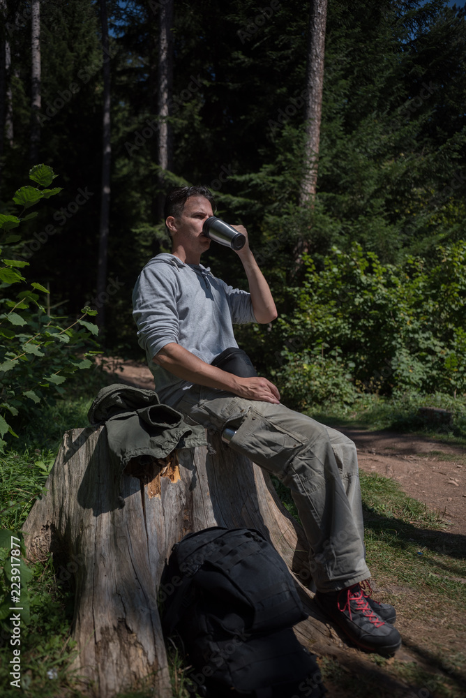 young man drinking from cup and resting on stump after a long hike 