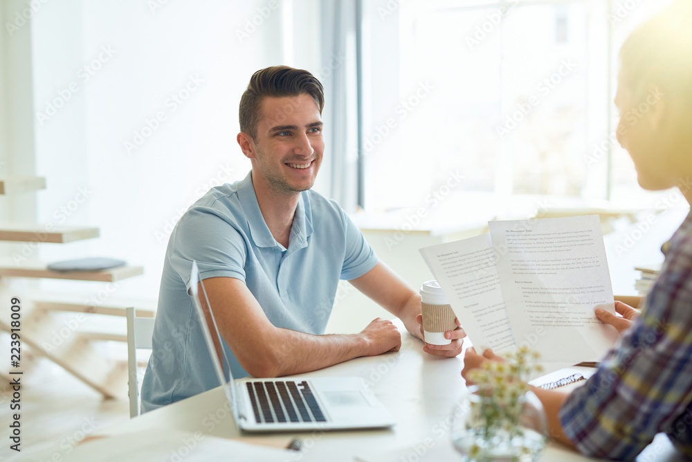 Smiling young businessman with drink looking at his colleague during discussion of papers Smiling young businessman with drink looking at his colleague during discussion of papers