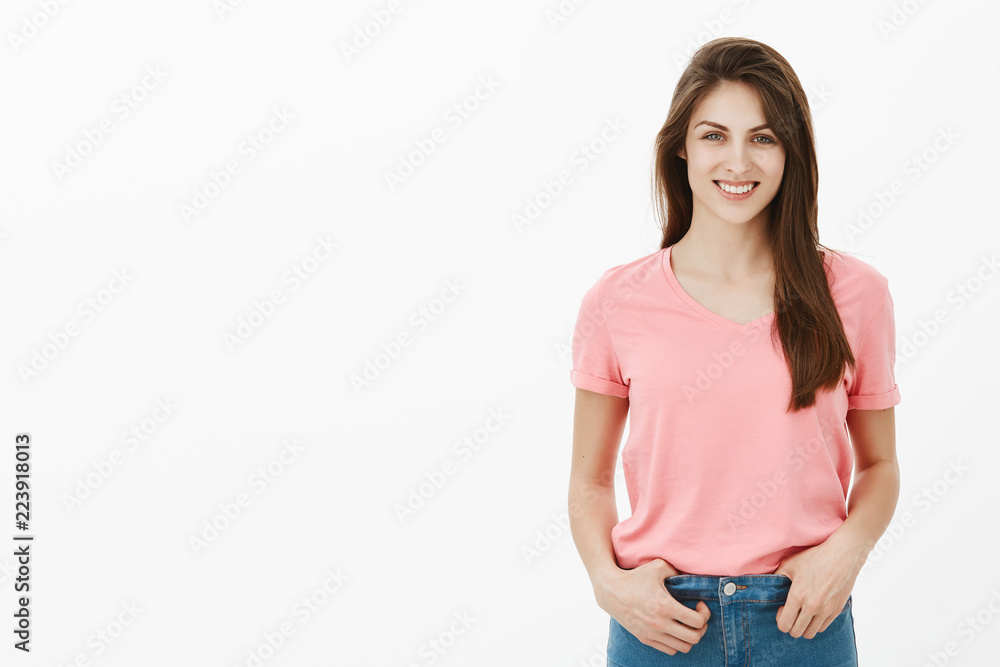 Girl gives interview about her success in science. Positive attractive brunette female student in pink t-shirt, holding hands on jeans and smiling broadly while talking casually over grey wall Girl gives interview about her success in science. Positive attractive brunette female student in pink t-shirt, holding hands on jeans and smiling broadly while talking casually over grey wall