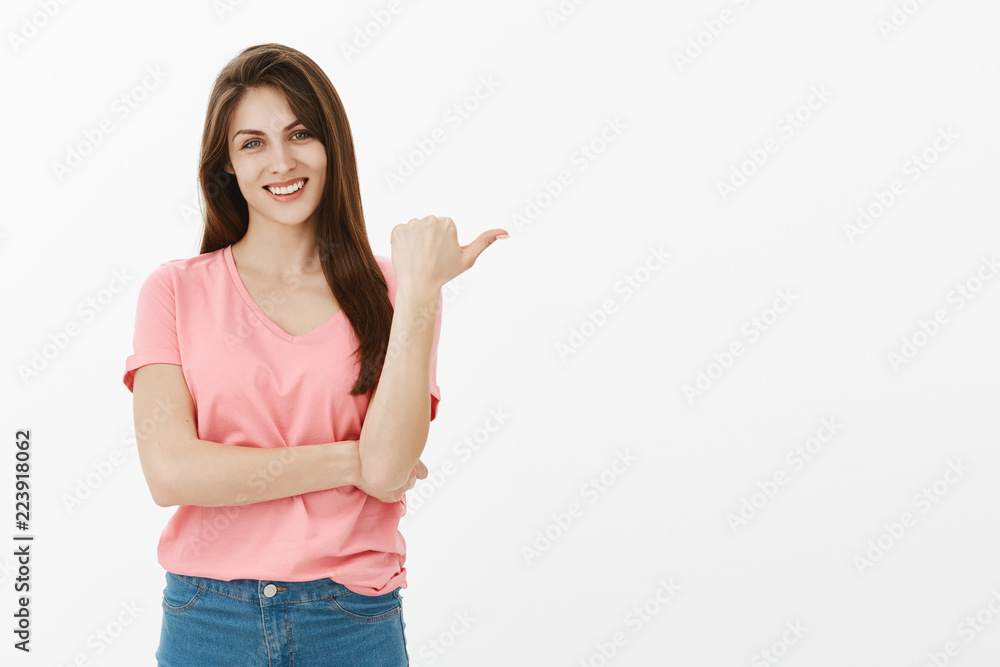 Portrait of proud happy european mom in pink t-shirt pointing right with thumb and smiling joyfully, directing at something impressing and curious, giving suggestion while standing over grey wall Portrait of proud happy european mom in pink t-shirt pointing right with thumb and smiling joyfully, directing at something impressing and curious, giving suggestion while standing over grey wall