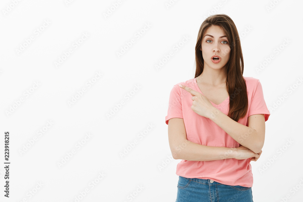 Girl asking advice while pointing at dress she wants to buy. Portrait of good-looking busy young woman in casual outfit, pointing at upper left corner and talking to camera over grey wall Girl asking advice while pointing at dress she wants to buy. Portrait of good-looking busy young woman in casual outfit, pointing at upper left corner and talking to camera over grey wall