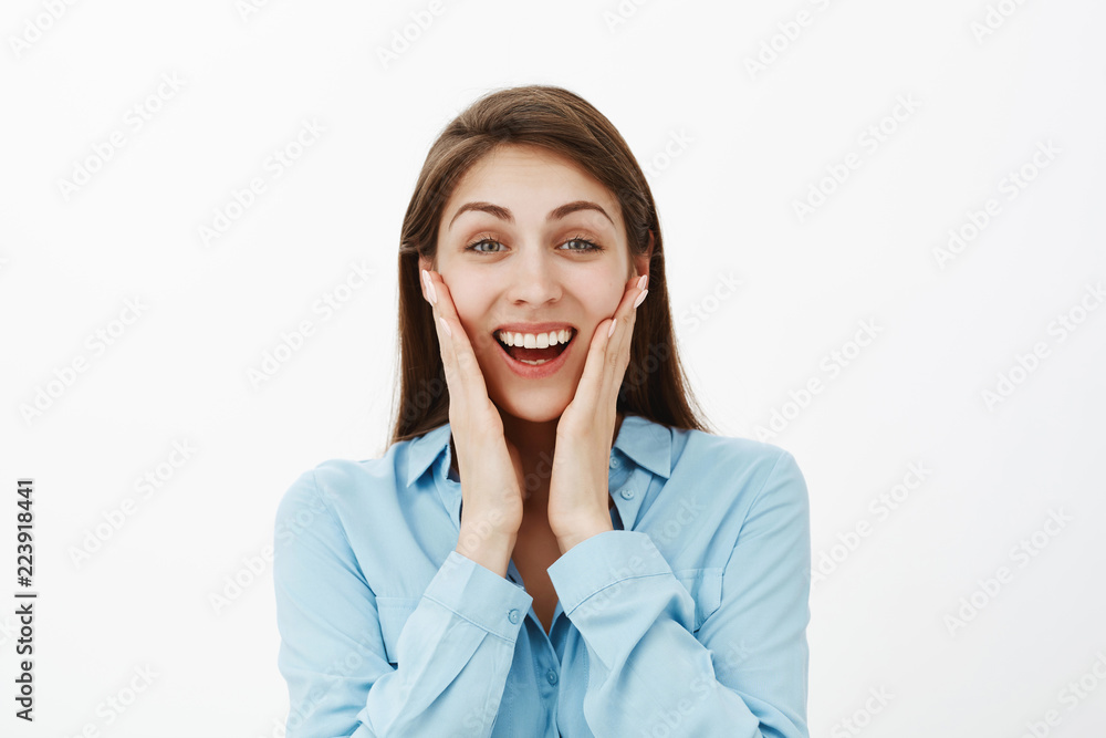 Close-up shot of satisfied surprised attractive woman with brown hair, touching cheeks with palms and smiling broadly, receiving great news or awesome gift, being impressed and amazed over grey wall Close-up shot of satisfied surprised attractive woman with brown hair, touching cheeks with palms and smiling broadly, receiving great news or awesome gift, being impressed and amazed over grey wall