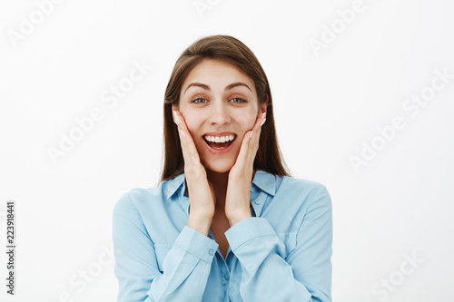 Close-up shot of satisfied surprised attractive woman with brown hair, touching cheeks with palms and smiling broadly, receiving great news or awesome gift, being impressed and amazed over grey wall