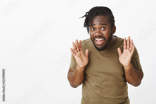 Uninvolved attractive african american guy with braid haircut raising palms in surrender and gazing timid at camera, apologizing or expressing unawareness, being confused and clueless over grey wall photo