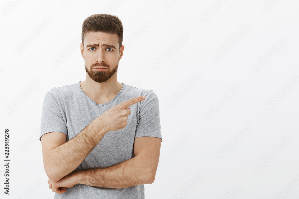 Sad and cute charming bearded male model in grey t-shirt frowning making gloomy face with frowned eyebrows, sulking pointing right expressing regret and envy standing unhappy over white background Sad and cute charming bearded male model in grey t-shirt frowning making gloomy face with frowned eyebrows, sulking pointing right expressing regret and envy standing unhappy over white background