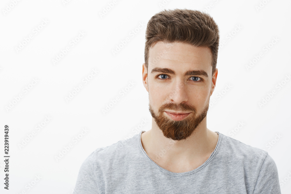 Fototapeta premium Waist-up shot of handsome sensual and confident young man with beard, moustache and blue eyes smiling looking sincere and self-assured at camera as with delighted smile against gray background