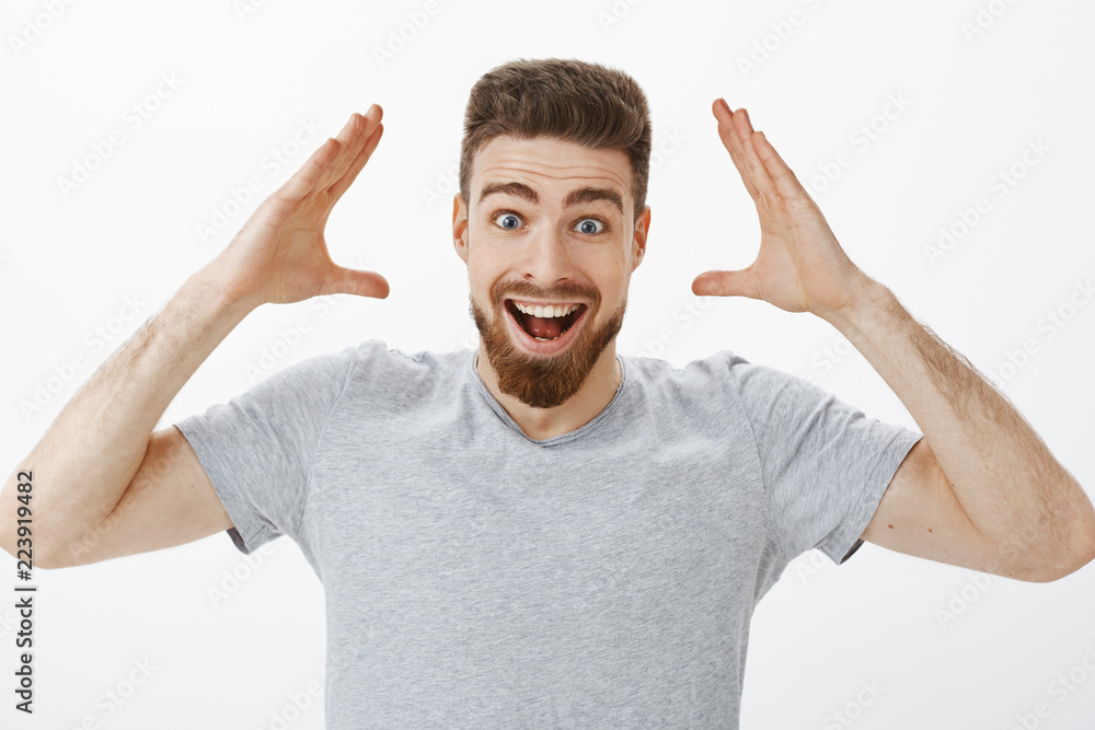 Can you imagine I got job. Charismatic joyful and excited handsome male model with beard in grey t-shirt raising hands near head smiling broadly receiving good news and retelling it friends happily Can you imagine I got job. Charismatic joyful and excited handsome male model with beard in grey t-shirt raising hands near head smiling broadly receiving good news and retelling it friends happily