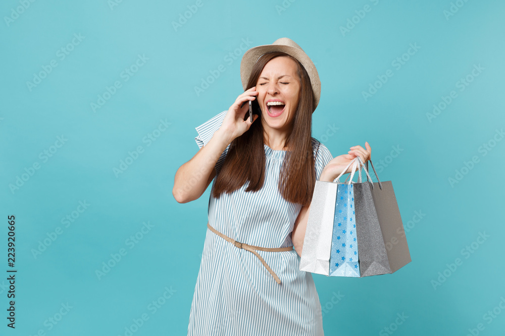 Portrait fun smiling woman in summer dress, straw hat holding packages bags with purchases after shopping, talking on mobile cellphone isolated on blue pastel background. Copy space for advertisement. Portrait fun smiling woman in summer dress, straw hat holding packages bags with purchases after shopping, talking on mobile cellphone isolated on blue pastel background. Copy space for advertisement.