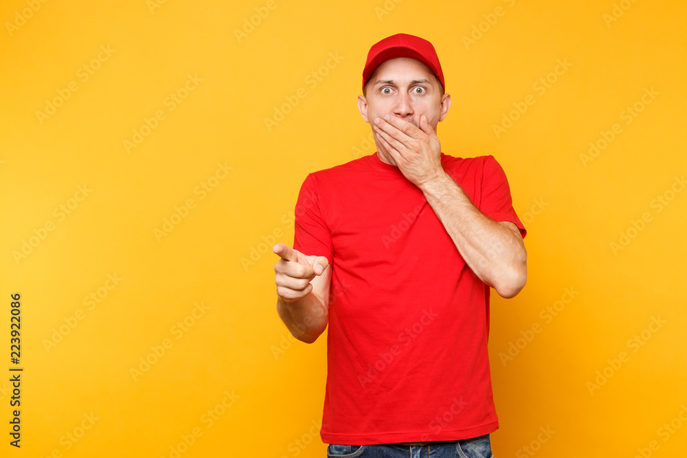 Delivery man in red uniform isolated on yellow orange background. Professional shocked male employee in cap, t-shirt working as courier or dealer cover mouth with hand. Service concept. Copy space. Delivery man in red uniform isolated on yellow orange background. Professional shocked male employee in cap, t-shirt working as courier or dealer cover mouth with hand. Service concept. Copy space.