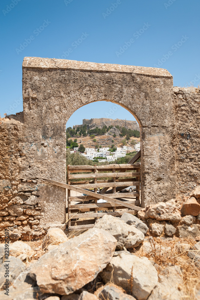 Distant view through stone arch opening at Lindos Town and Castle with ancient ruins of the Acropolis on sunny warm day. Island of Rhodes, Greece.