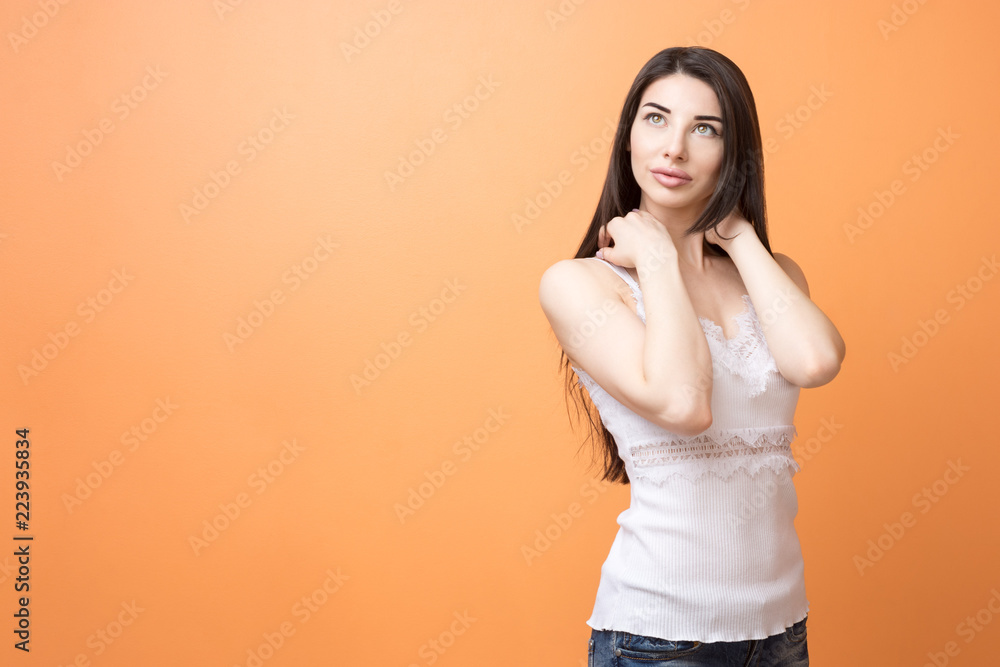 Portrait of a young beautiful brunette pensive woman with long hair holding her hand near her head and looking up, thinking or dreaming about something against orange background. Portrait of a young beautiful brunette pensive woman with long hair holding her hand near her head and looking up, thinking or dreaming about something against orange background.