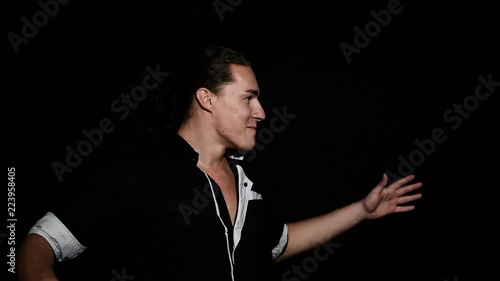 Young attractive stylish brown eyed man with dark curly hair dressed in black shirt with short sleeves and trousers is playfully smiling, speaking and shows dance movements in front of camera.
