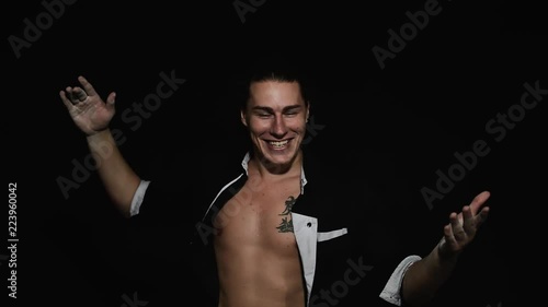 Handsome athletic young brown eyed guy in unbuttoned shirt with tattoo on chest is smiling and throwing white powder on himself in front of camera on black matte background. photo