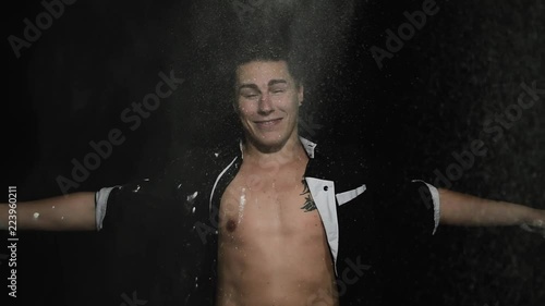 Handsome athletic young brown eyed male in unbuttoned shirt with tattoo on chest is smiling and dramatically throwing white powder on himself in front of camera on black matte background. photo