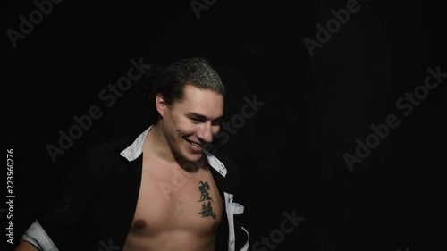 Handsome athletic young brown eyed male in unbuttoned shirt with tattoo on chest is smiling and dramatically throwing white powder around himself in front of camera on black matte background. photo