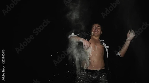 White powder is being thrown on handsome, sporty young brown eyed guy with dark hair combed into bundle and wearing unbuttoned shirt with tattoo on chest in front of camera on black matte background photo