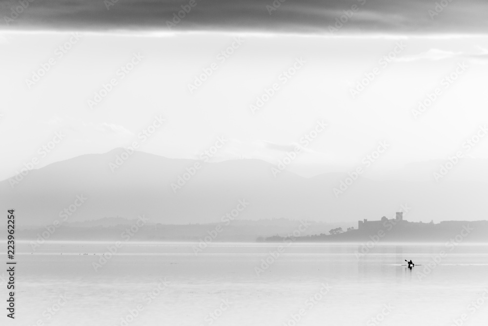 Beautiful view of Trasimeno lake at sunset with birds on water, a man on a canoe and Castiglione del Lago town in the background