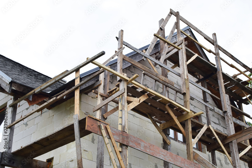 Wooden scaffolding next to white residental house built of aerated concrete blocks with grey asphalt shingles roof on sky background with copy space. House improvement process nd DIY concept. Wooden scaffolding next to white residental house built of aerated concrete blocks with grey asphalt shingles roof on sky background with copy space. House improvement process nd DIY concept.