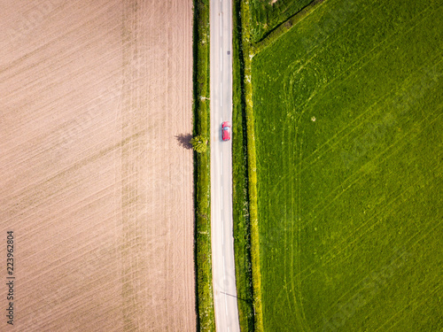 Aerial view looking down on a rural road in the UK countryside with a car racing along it. On a bright sunny day, farmland and crops can be seen either side of the road