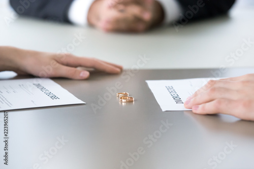 Close up of marriage rings lying on table along with divorce decrees and couple hands, husband and wife sign paper separating or breaking up officially, spouses end relationships at lawyer office photo