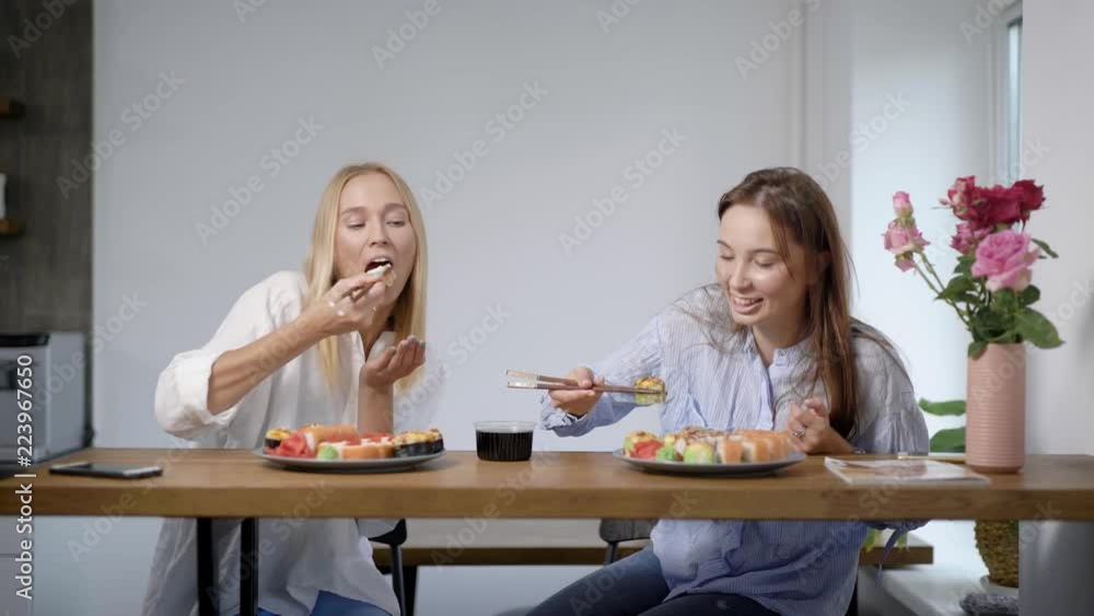 Two lovely young girls eating sushi rolls and talking, friends together in kitchen.