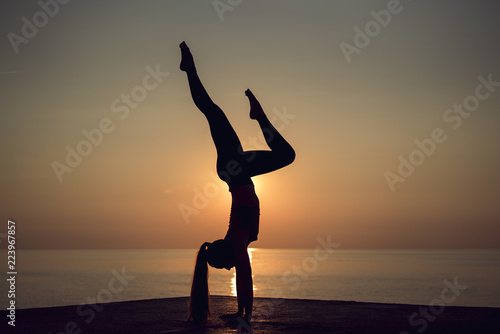 Silhouette of young gymnast standing on hands in a beautiful pose on the beach at sunset. Woman doing exercise on the pier. Professional yoga teacher practicing handstanding outdoors. Powerful woman. photo