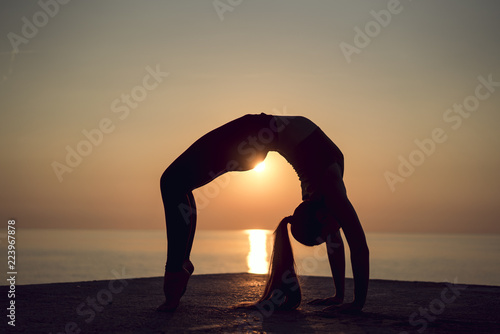 Silhouette of young gymnast doing a wheel pose on the beach at sunset. Woman practicing yoga or pilates on the pier. Bridge Pose, Urdhva Dhanurasana (Upward Bow), Chakrasana (Wheel). Side view.