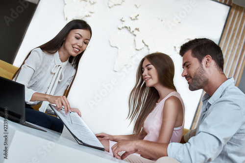 Young lovely couple sitting with manager at the travel agency office and reviewing contract for a summer vacation
