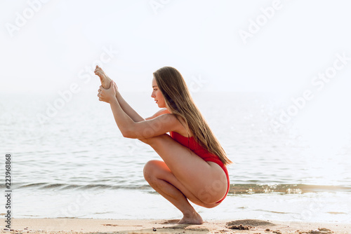 Young woman practicing yoga, stretching legs to be flexible and fit. Healthy girl doing exercises on the beach on the sunrise. Relaxing early in the morning. Concept of calmless and health. photo