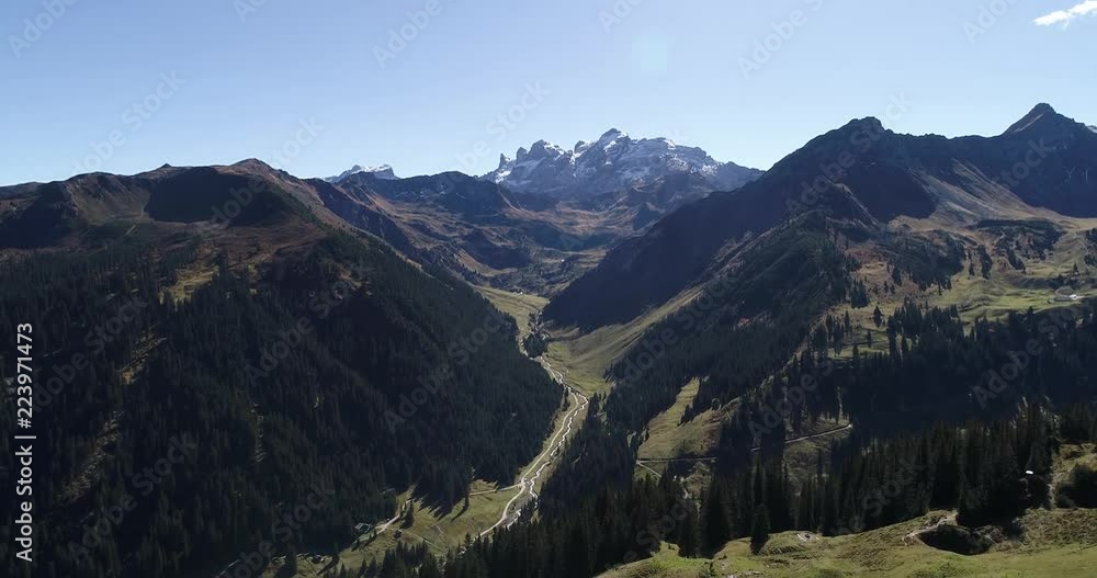 Scenic aerial scene. River between two mountains and snowy summit peak behind sunny summer day. Austria alps. Drone rising up and filming green forests and clear blue sky during midday. Scenic aerial scene. River between two mountains and snowy summit peak behind sunny summer day. Austria alps. Drone rising up and filming green forests and clear blue sky during midday.