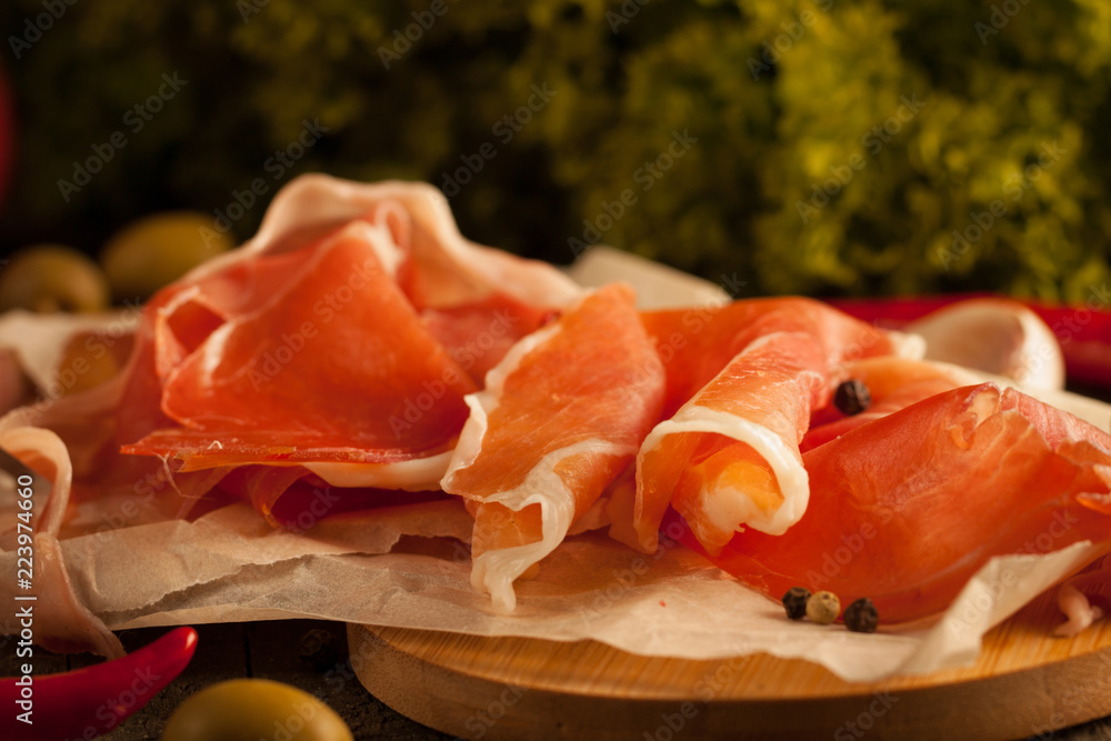 Closeup macro photo of thin slices of prosciutto with green olives, spices and food background on wooden cutting board. Raw dried meat. Closeup macro photo of thin slices of prosciutto with green olives, spices and food background on wooden cutting board. Raw dried meat.