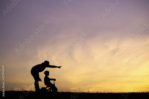 The silhouette photo of mother and her daughter riding bicycle in the park with sweet sky at sunset.