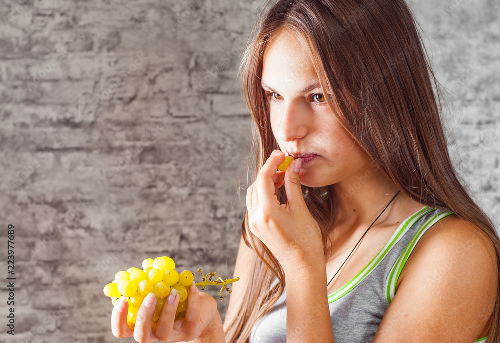 portrait of young teenager brunette girl with long hair eating grape on gray wall background